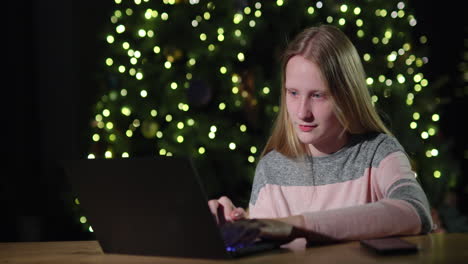 Ein-Junges-Mädchen-Benutzt-Einen-Laptop-Vor-Dem-Hintergrund-Eines-Weihnachtsbaums