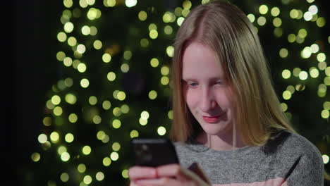 A-teenage-girl-uses-a-smartphone-on-the-background-of-a-Christmas-tree