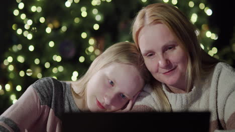 Mom-and-daughter-are-looking-at-the-laptop-screen-together.-Sitting-in-front-of-the-blurred-lights-of-the-Christmas-tree