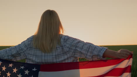 Freihandaufnahme:-Eine-Frau-Mit-Einer-Amerikanischen-Flagge-Auf-Den-Schultern-Sieht-Zu,-Wie-Die-Sonne-über-Einem-Weizenfeld-Untergeht.