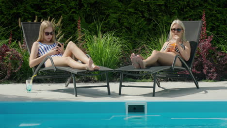 A-woman-and-her-daughter-are-relaxing-on-sun-loungers-by-the-pool,-drinking-soft-drinks