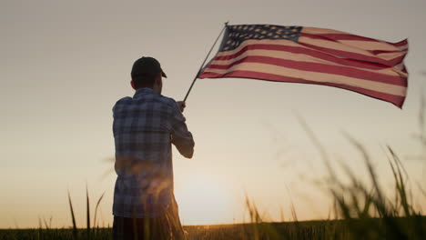 Silhouette-Eines-Jungen-Mannes,-Der-Die-Amerikanische-Flagge-Schwenkt.-Bei-Sonnenuntergang-Auf-Einem-Weizenfeld-Stehen