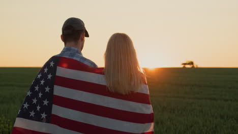 A-couple-of-farmers-with-an-American-flag-on-their-shoulders-look-at-a-wheat-field-where-a-tractor-is-working-in-the-distance.
