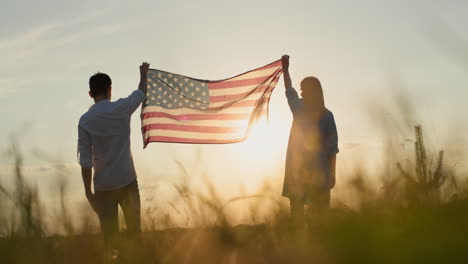 Man-and-woman-raising-the-US-flag-over-a-field-of-wheat-at-sunset