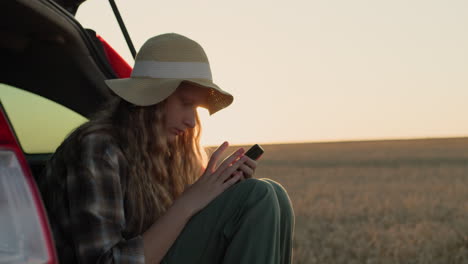 A-teenage-girl-sits-in-the-trunk-of-a-car,-uses-a-smartphone.-Against-the-backdrop-of-a-rural-landscape-where-the-sun-sets