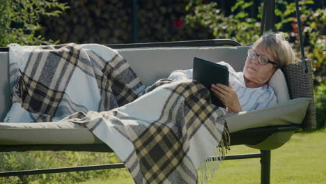 An-elderly-woman-lies-under-a-blanket-in-a-garden-swing,-using-a-tablet.-Relaxing-in-the-backyard-of-the-house