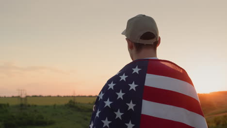 A-man-with-an-American-flag-on-his-shoulders-admires-the-sunrise-over-a-picturesque-valley.-Independence-Day-concept
