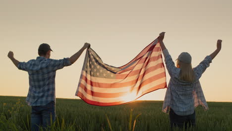 Ein-Mann-Und-Eine-Frau-Hissen-Die-US-Flagge-über-Einem-Weizenfeld,-Während-Die-Sonne-Untergeht.-Konzept-Zum-Unabhängigkeitstag-Der-USA
