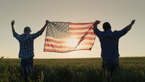 Two-men-raise-up-the-US-flag-against-the-backdrop-of-a-wheat-field-at-sunset