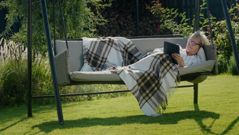 Happy-senior-woman-lies-under-a-blanket-in-a-garden-swing,-using-a-tablet.-Relaxing-in-the-backyard-of-the-house
