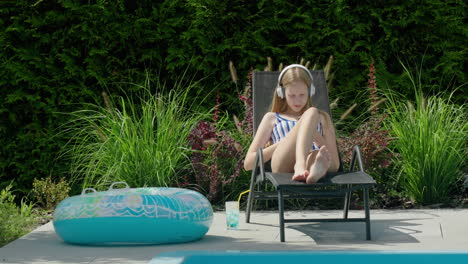 A-teenage-girl-is-resting-in-a-sun-lounger-by-the-pool.-Uses-a-smartphone,-listens-to-music-on-headphones