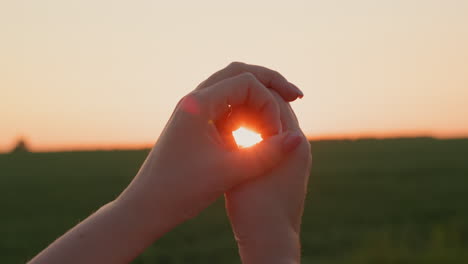 Women's-hands-hold-the-disk-of-the-sun,-which-sets-over-a-field-of-wheat