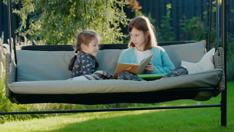 Girl-reading-a-book-to-her-younger-sister,-relaxing-on-a-swing-in-the-backyard-of-the-house