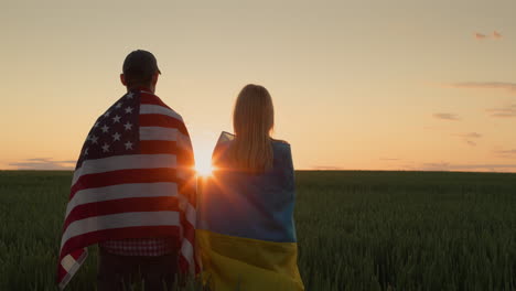 Happy-couple-with-the-flags-of-Ukraine-and-the-USA-stand-side-by-side-and-look-at-the-sunrise-over-a-field-of-wheat