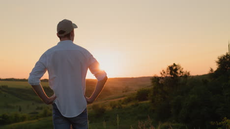A-confident-young-farmer-stands-in-front-of-a-picturesque-countryside-as-the-sun-sets.-View-from-the-back