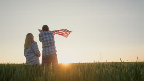An-energetic-couple-waving-the-American-flag-in-front-of-a-field-of-wheat-as-the-sun-sets.-4th-of-july-concept