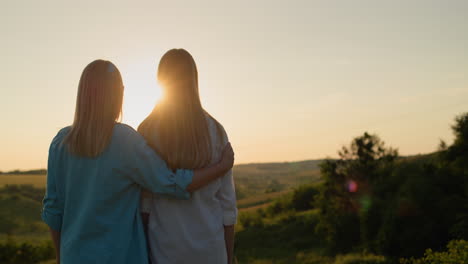 A-woman-hugs-her-teenage-daughter-and-together-they-watch-the-sun-go-down-over-a-picturesque-valley