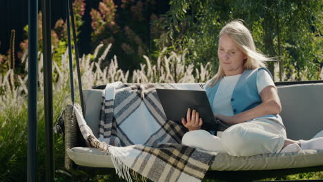A-young-woman-uses-a-laptop.-Resting-in-a-garden-swing-in-the-backyard-of-a-house