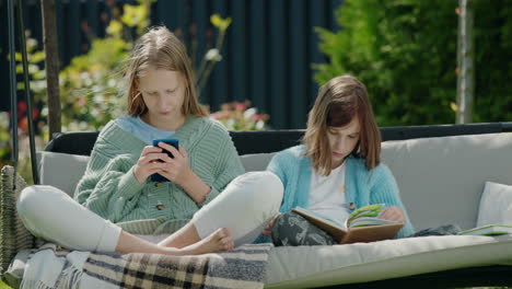 Zwei-Freunde-Entspannen-Sich-Auf-Einer-Gartenschaukel.-Ein-Mädchen-Benutzt-Ein-Smartphone,-Ein-Anderes-Liest-Ein-Buch