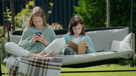 Two-teen-girls-are-relaxing-on-a-garden-swing.-One-girl-uses-a-smartphone,-another-reads-a-book