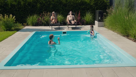 Die-Familie-Entspannt-An-Ihrem-Pool-Im-Hinterhof-Des-Hauses.-Erwachsene-Sonnen-Sich-Auf-Sonnenliegen,-Zwei-Kinder-Schwimmen-Im-Pool