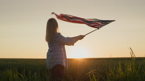 A-young-woman-waving-the-usa-flag,-stands-in-the-background-of-a-picturesque-field-of-wheat-where-the-sun-sets.-Slow-motion-video