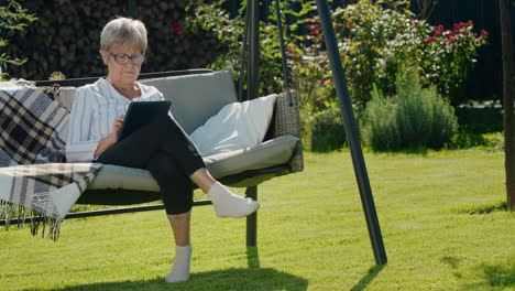 Portrait-of-senior-woman-using-a-tablet.-Sitting-in-a-garden-swing-in-the-backyard-of-a-house