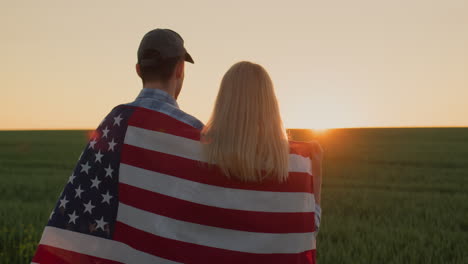 Junges-Paar-Mit-USA-Flagge-Auf-Den-Schultern,-Das-Den-Sonnenaufgang-über-Dem-Weizenfeld-Bewundert