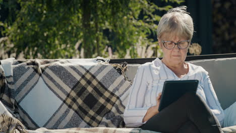 Portrait-of-an-elderly-woman-using-a-tablet.-Sitting-in-a-garden-swing-in-the-backyard-of-a-house
