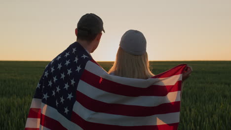Junges-Paar-Mit-Amerikanischer-Flagge-Auf-Den-Schultern-Freut-Sich-Im-Weizenfeld-Bei-Sonnenuntergang