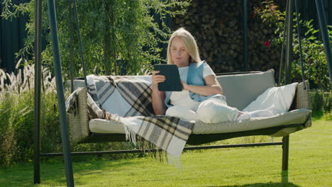 Cute-woman-uses-a-tablet.-Resting-in-a-garden-swing-in-the-backyard-of-a-house