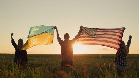 A-group-of-friends-raises-the-flag-of-the-United-States-and-the-flag-of-Ukraine-over-their-heads.-Standing-against-the-backdrop-of-a-field-of-wheat-where-the-sun-sets