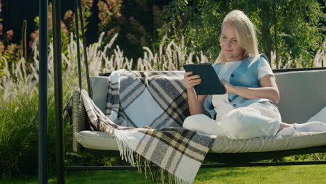 Happy-woman-uses-a-tablet.-Resting-in-a-garden-swing-in-the-backyard-of-a-house.-4k-video