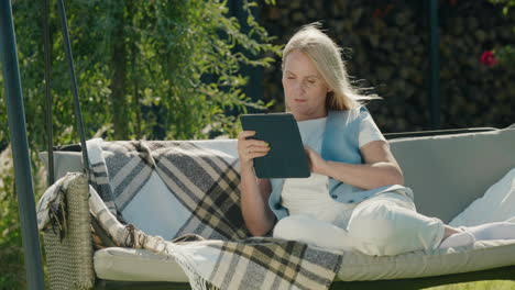 Happy-woman-uses-a-tablet.-Resting-in-a-garden-swing-in-the-backyard-of-a-house