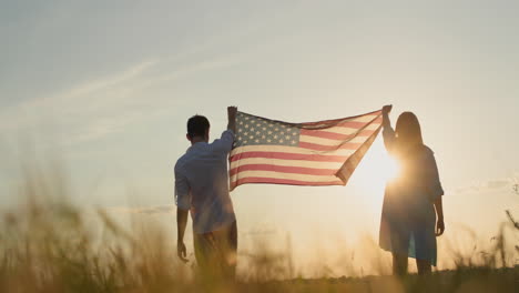 Ein-Paar-Hisst-Die-US-Flagge-über-Einem-Weizenfeld,-Während-Die-Sonne-Untergeht.-Konzept-Zum-Unabhängigkeitstag-Der-USA.-4K-Video