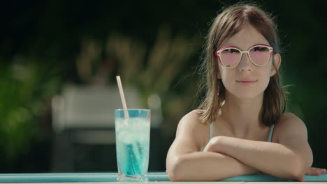 Portrait-of-a-cute-little-girl-in-sunglasses-by-the-side-of-the-pool.-Nearby-is-a-cool-cocktail