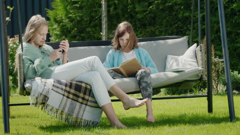 Zwei-Freunde-Entspannen-Sich-Auf-Einer-Gartenschaukel.-Ein-Mädchen-Benutzt-Ein-Smartphone,-Ein-Anderes-Liest-Ein-Buch