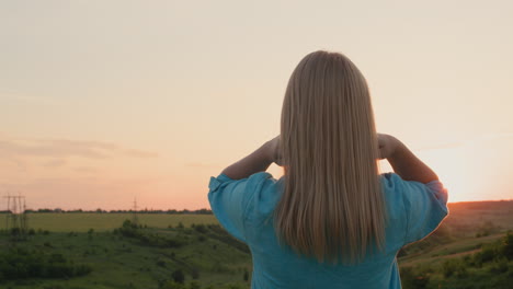 A-woman-straightens-her-hair,-stands-against-the-backdrop-of-a-picturesque-landscape-where-the-sun-sets