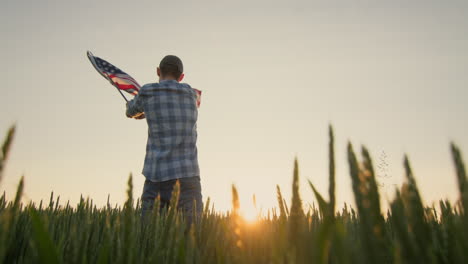 American-farmer-waving-the-US-flag-against-the-backdrop-of-a-field-of-wheat-where-the-sun-rises.-Low-angle-shot