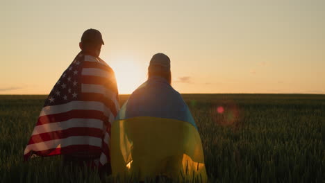 Couple-with-the-flags-of-Ukraine-and-the-USA-stand-side-by-side-and-look-at-the-sunset-over-a-field-of-wheat