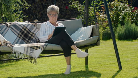 Portrait-of-senior-woman-using-a-digital-tablet.-Sitting-in-a-garden-swing-in-the-backyard-of-a-house