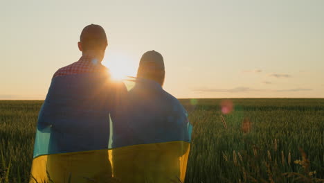 Ein-Junges-Paar-Mit-Der-Flagge-Der-Ukraine-Auf-Den-Schultern-Blickt-Auf-Den-Sonnenaufgang-über-Einem-Weizenfeld.-Konzept-Der-Hoffnung-Und-Des-Optimismus