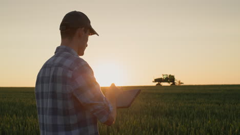 Successful-young-farmer-is-working-in-the-field.-A-tractor-is-driving-in-the-background.-A-man-uses-a-tablet---new-technologies-in-agriculture
