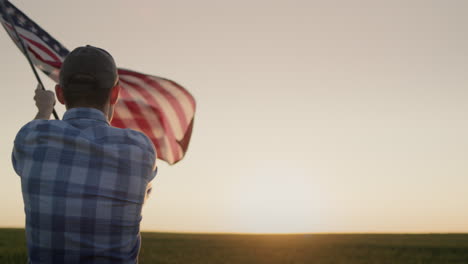 Silhouette-Eines-Jungen-Mannes,-Der-Die-Amerikanische-Flagge-Schwenkt.-Bei-Sonnenuntergang-Auf-Einem-Weizenfeld-Stehen.-Rückansicht