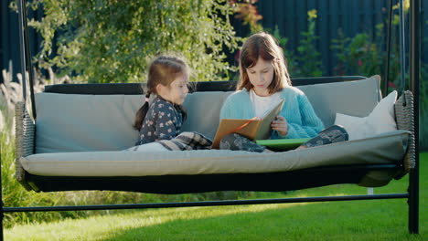 Girl-reading-a-book-to-her-younger-sister,-relaxing-on-a-swing-in-the-backyard-of-the-house