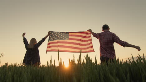 A-happy-couple-raises-up-the-US-flag,-standing-against-the-backdrop-of-a-wheat-field-at-sunset.-Low-angle-shot