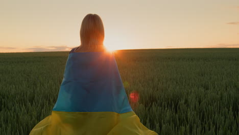 Una-Mujer-Con-La-Bandera-De-Ucrania-Sobre-Sus-Hombros-Mira-El-Amanecer-Sobre-Un-Campo-De-Trigo.-Soporte-Con-El-Concepto-De-Ucrania