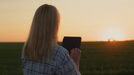 A-farmer-woman-uses-a-tablet.-Standing-in-a-field-where-a-tractor-is-working-in-the-distance-and-the-sun-is-setting