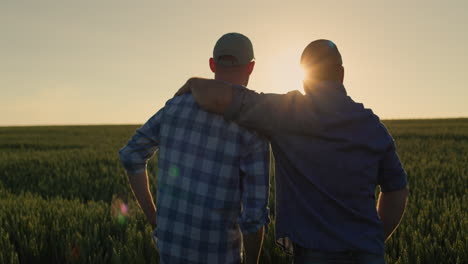 The-farmer-hugs-his-adult-son-and-looks-forward-together-to-the-wheat-field-where-the-sun-is-setting.-Family-business-and-traditions-concept
