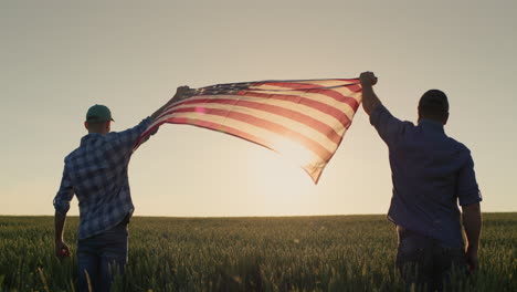 Zwei-Männer-Hissen-Die-US-Flagge-Vor-Dem-Hintergrund-Eines-Weizenfeldes-Bei-Sonnenuntergang